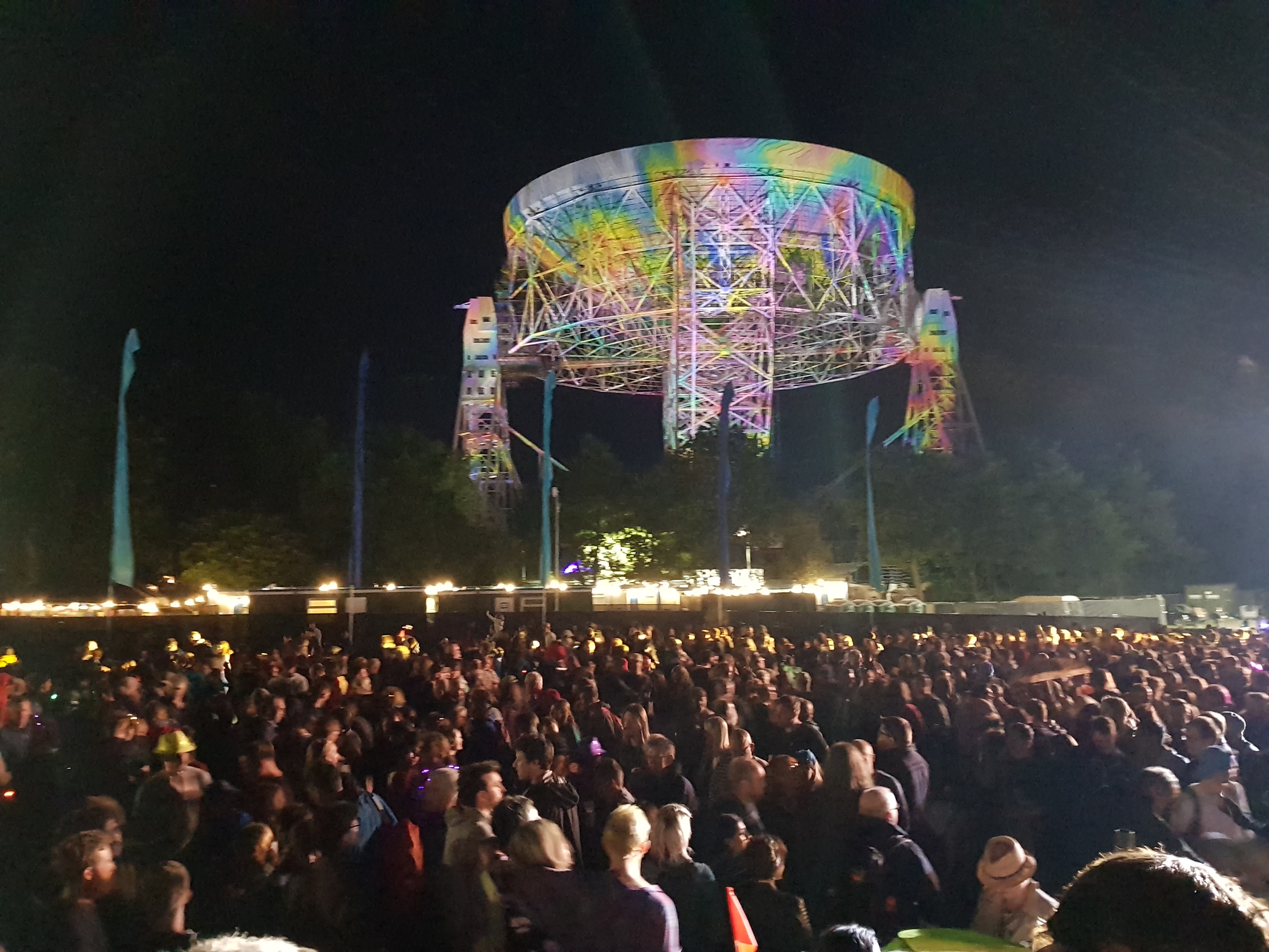 Lots of people standing in the foreground with a large radiotelescope dish in the background illuminated by multicoloured lights