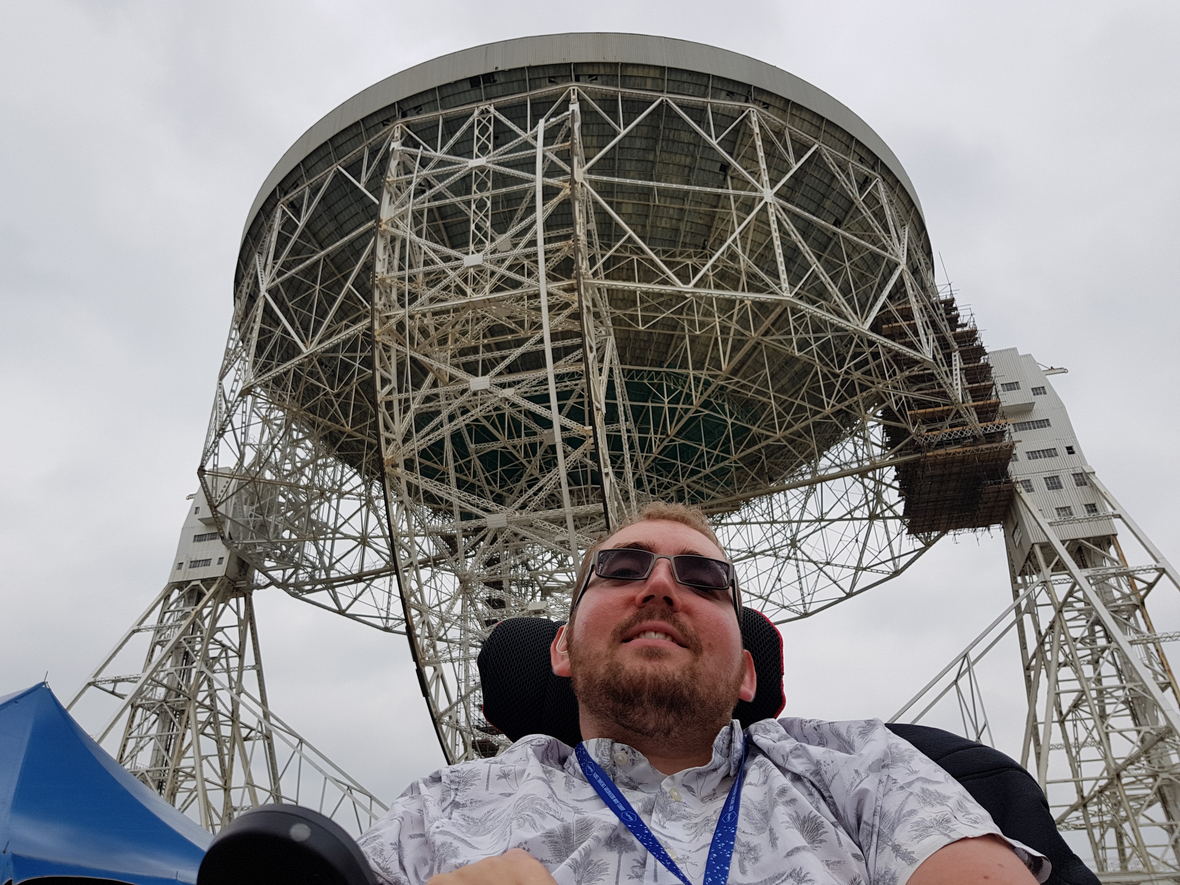 A bearded young man smiles at the camera with a big radiotelescope dish
