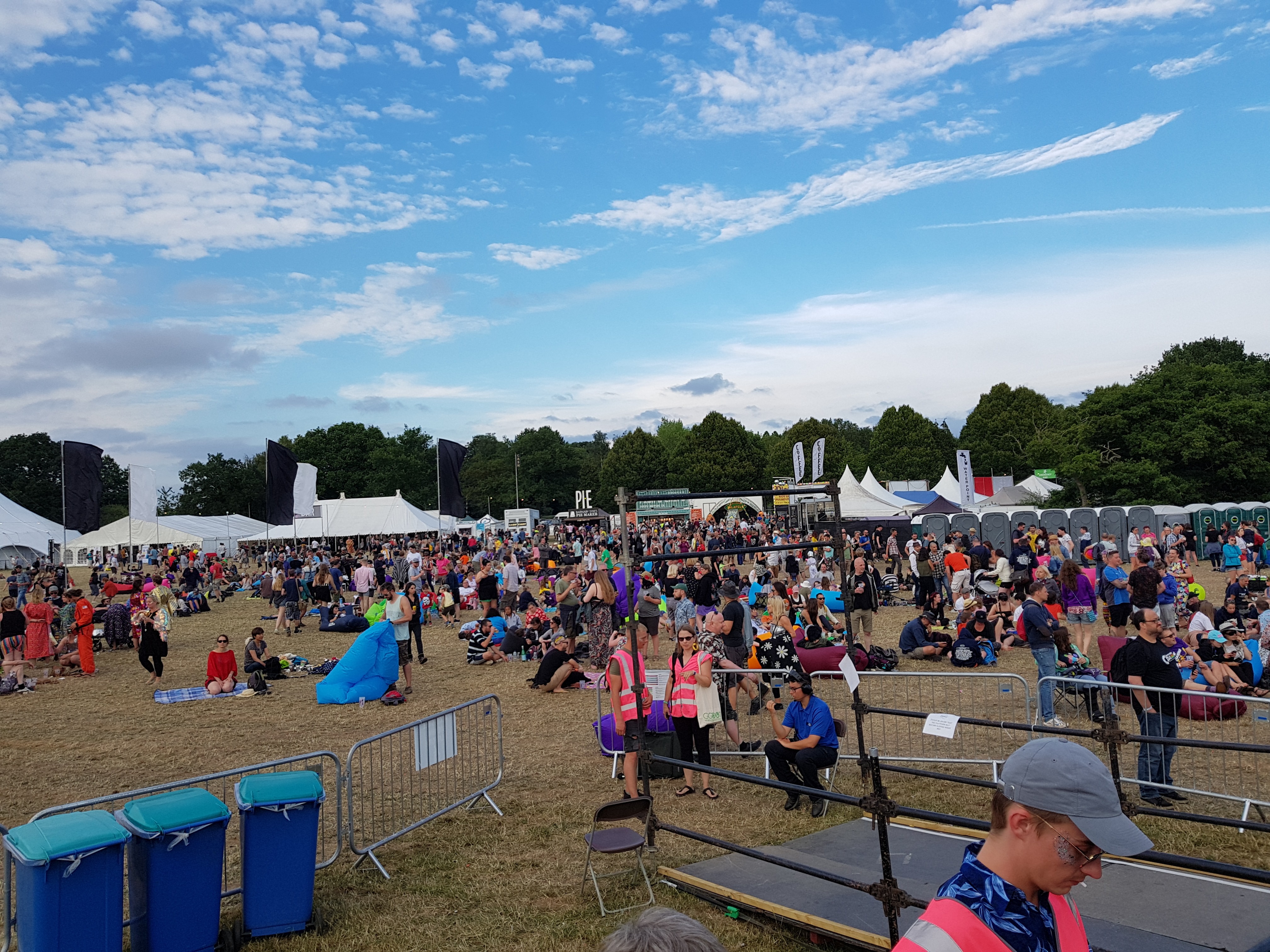 Lots of people are standing on the grass, some are sat on blankets or inflatable cushions. Some tents are in the background where DJs played