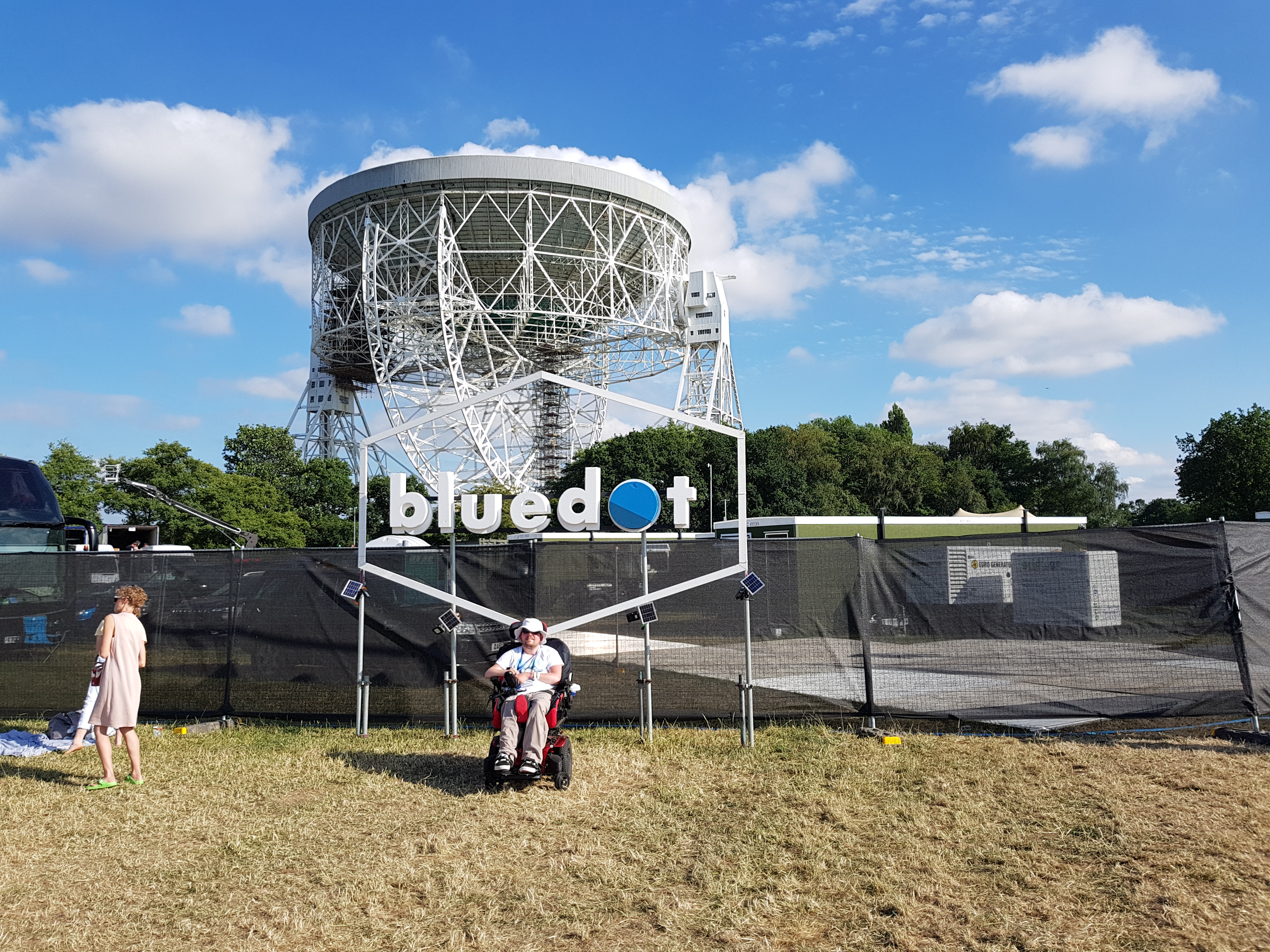 A young man in a Powerchair sitting in front of a sign that says "bluedot" with the huge Lovell radio telescope dish in the background.