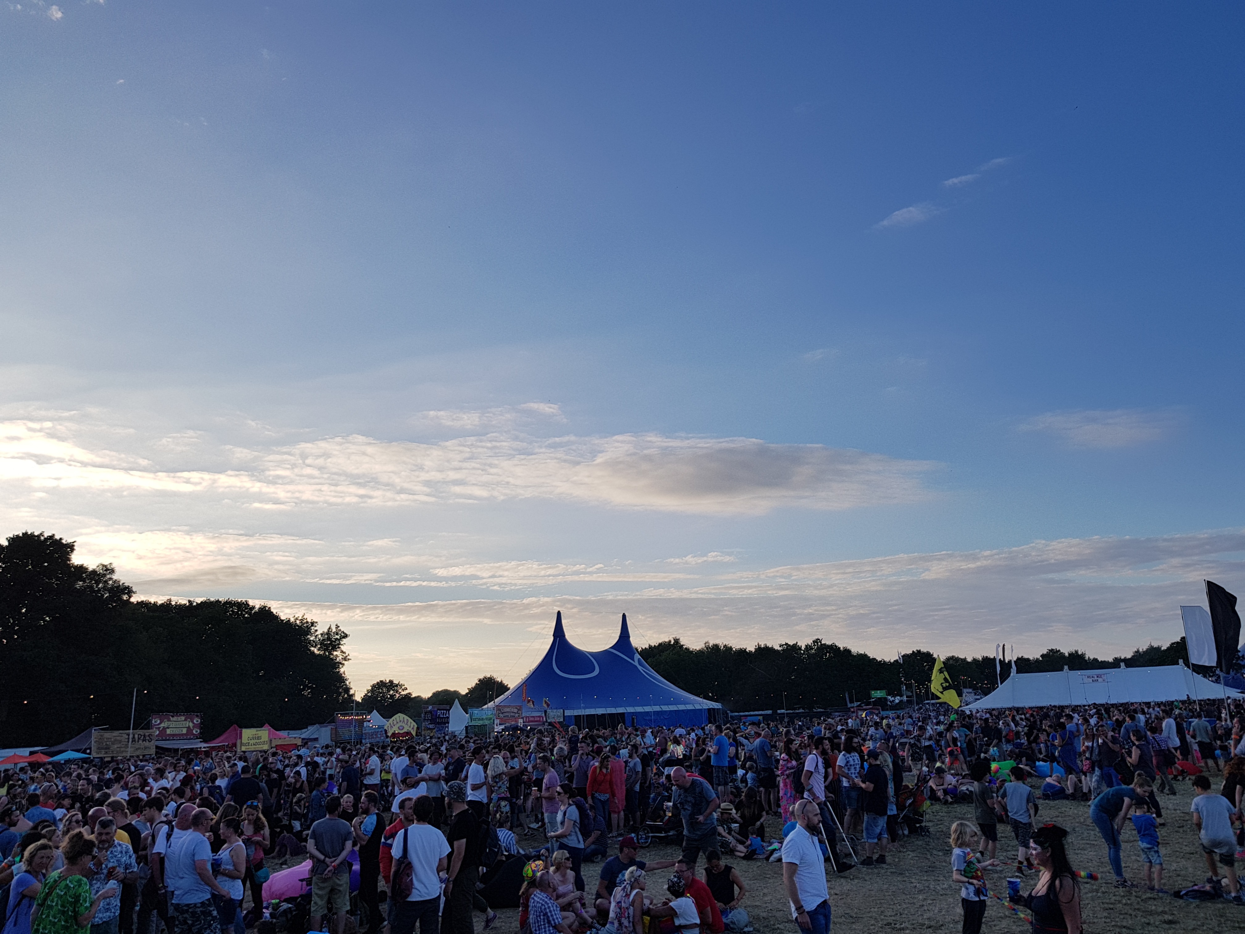 Lots of people milling about on the grass with a couple of marquees in the background, beneath a blue sky