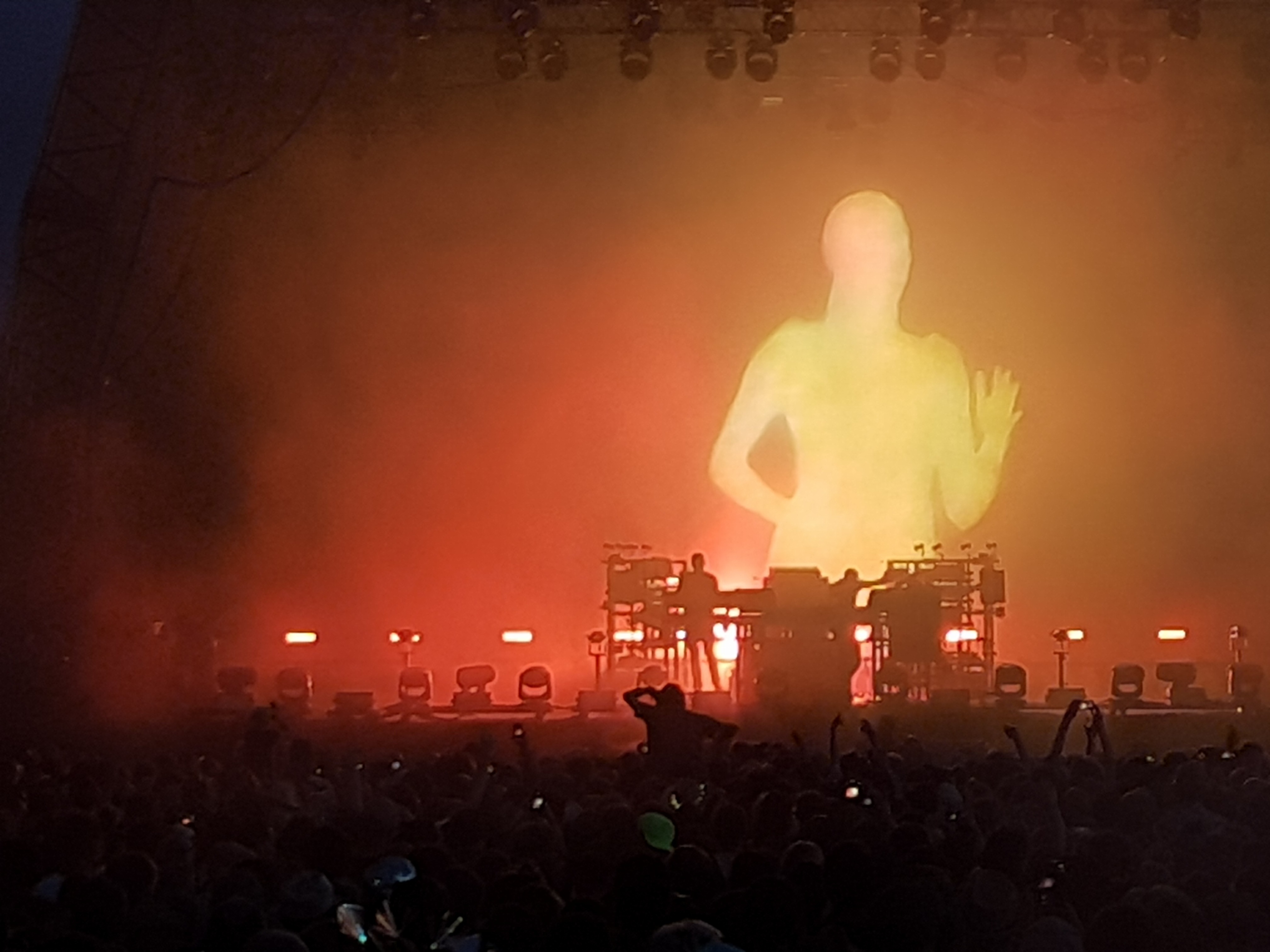 A close-up of the chemical Brothers onstage performing. They are standing behind the decks as a Large silhouette of a woman running is projected onto the screen behind them.