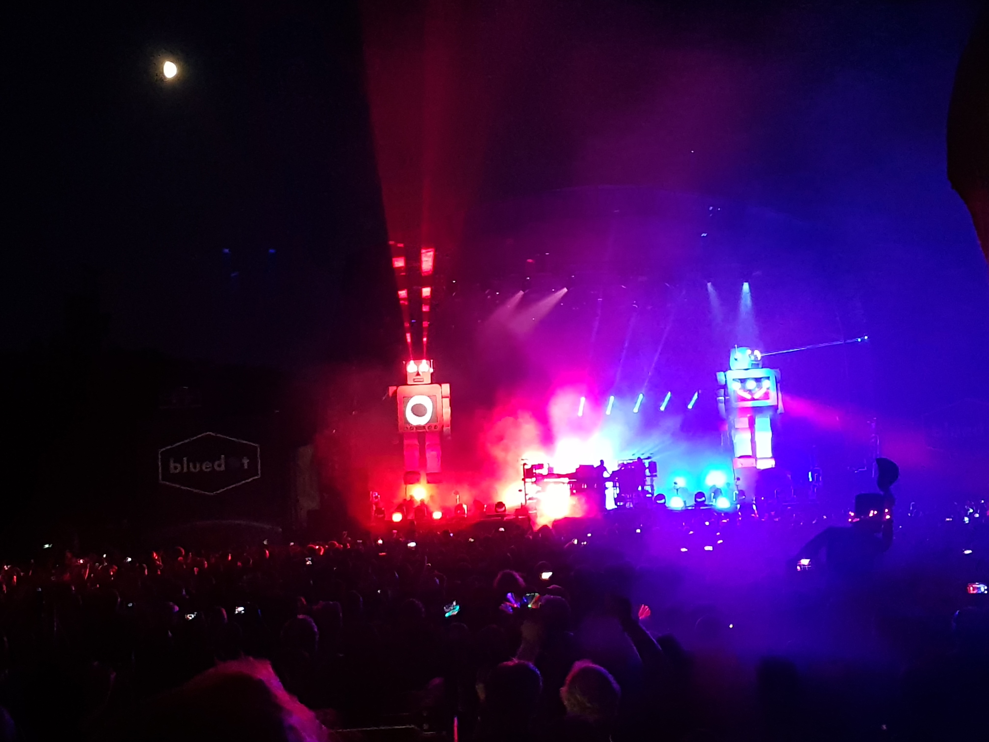 Lots of people standing in front of the stage at night as they hold up their phones to record the performance. Two Animatronic robots flank the stage with lasers coming out of their eyes