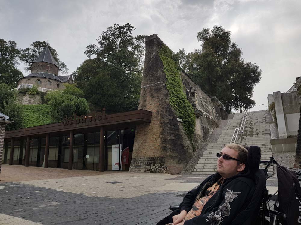 A person in a wheelchair is sitting in front of a building with modern architecture, featuring large glass windows and a wooden overhang. In the background, there is a historical structure with a conical roof, surrounded by greenery and stone walls.
