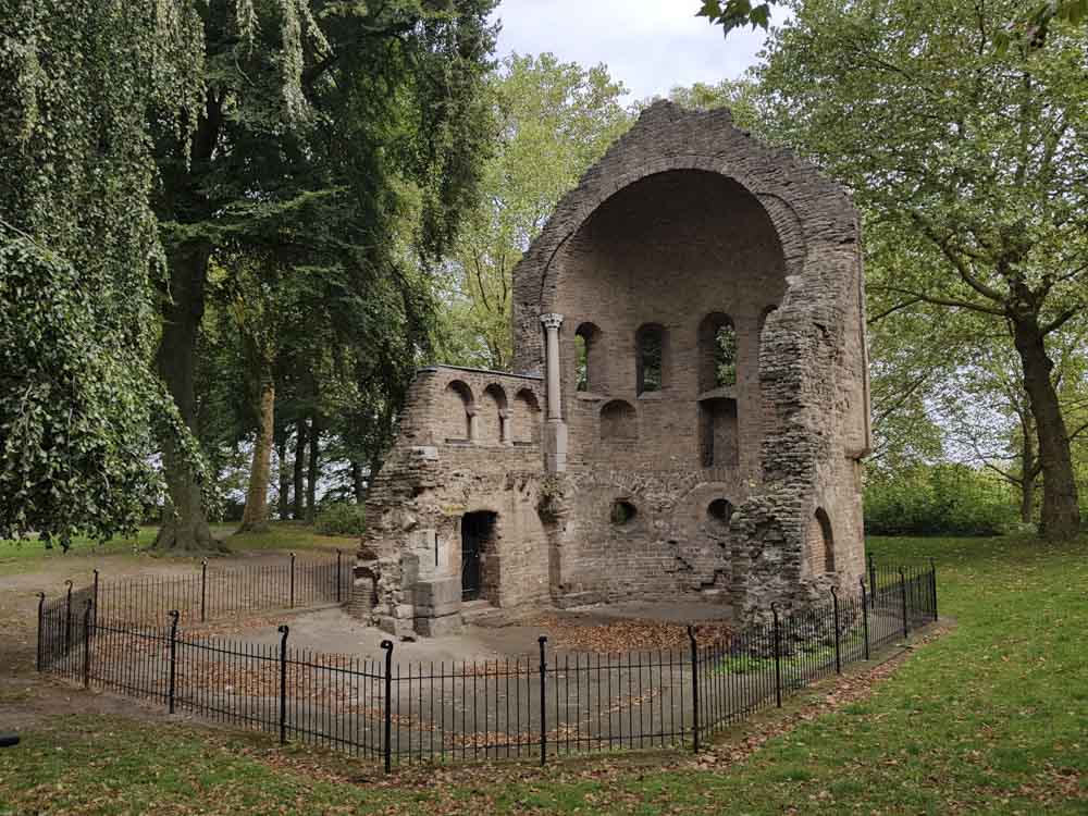 Ruins of a historic stone structure surrounded by trees and a fenced area, with leaves scattered on the ground.