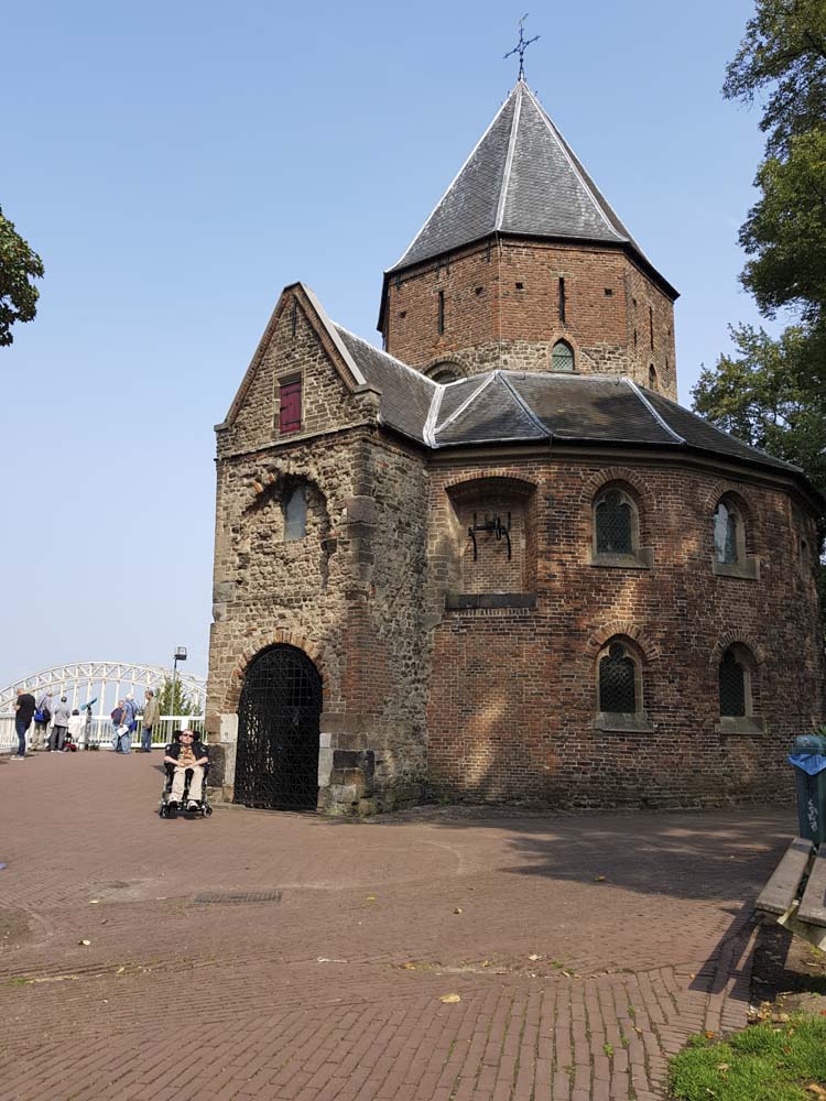 Historic stone building with a conical roof, located near a pedestrian area with people in the background.