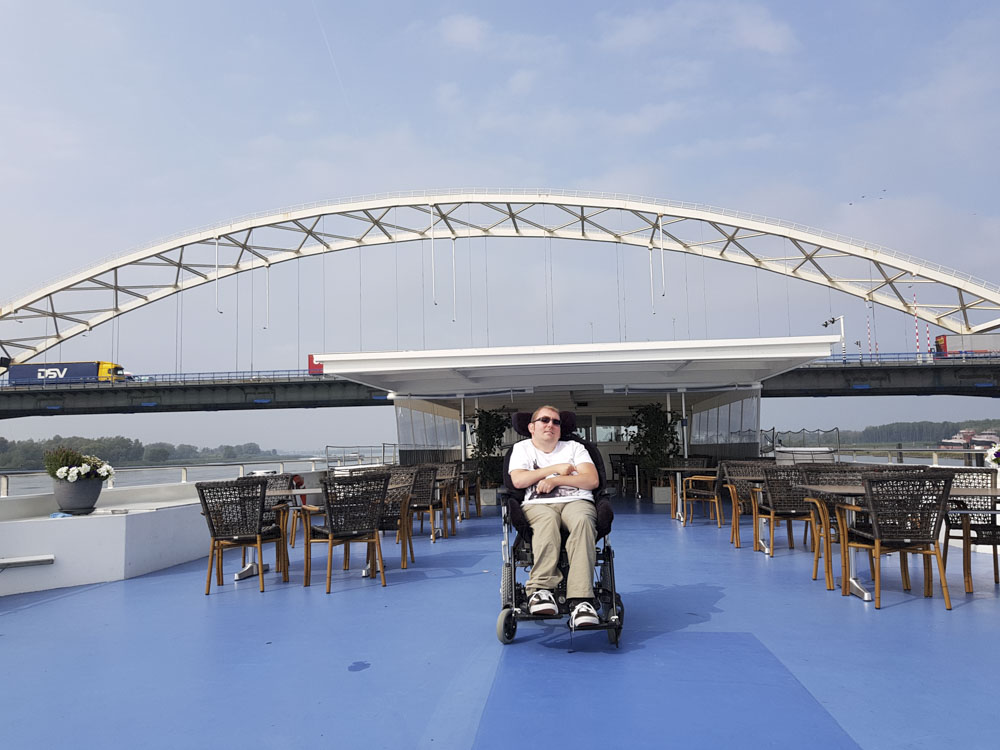 A person sitting in a wheelchair on the deck of a boat with a bridge overhead and outdoor seating in the background.