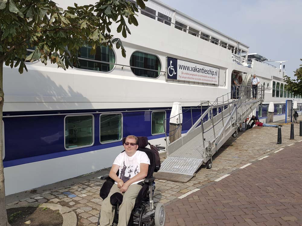 A person in a wheelchair sits in front of the Prins Willem Alexander ship, which features an accessible ramp. The ship is docked along a cobblestone path lined with trees.