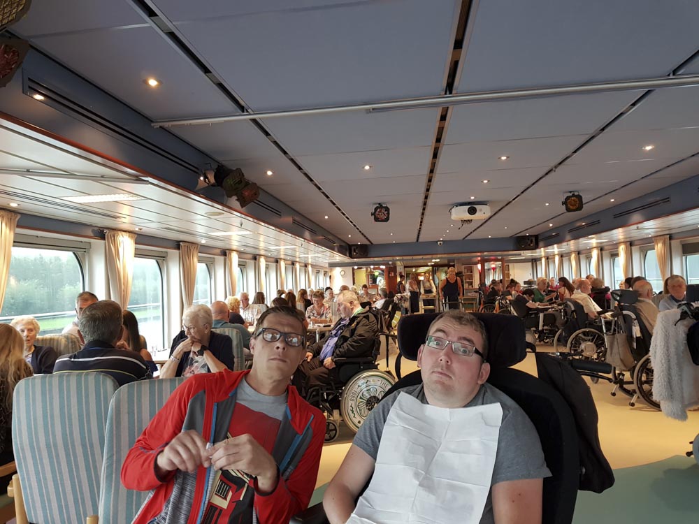 Interior view of a dining area on a canal cruise ship, featuring a mix of guests, some in wheelchairs, enjoying the atmosphere.