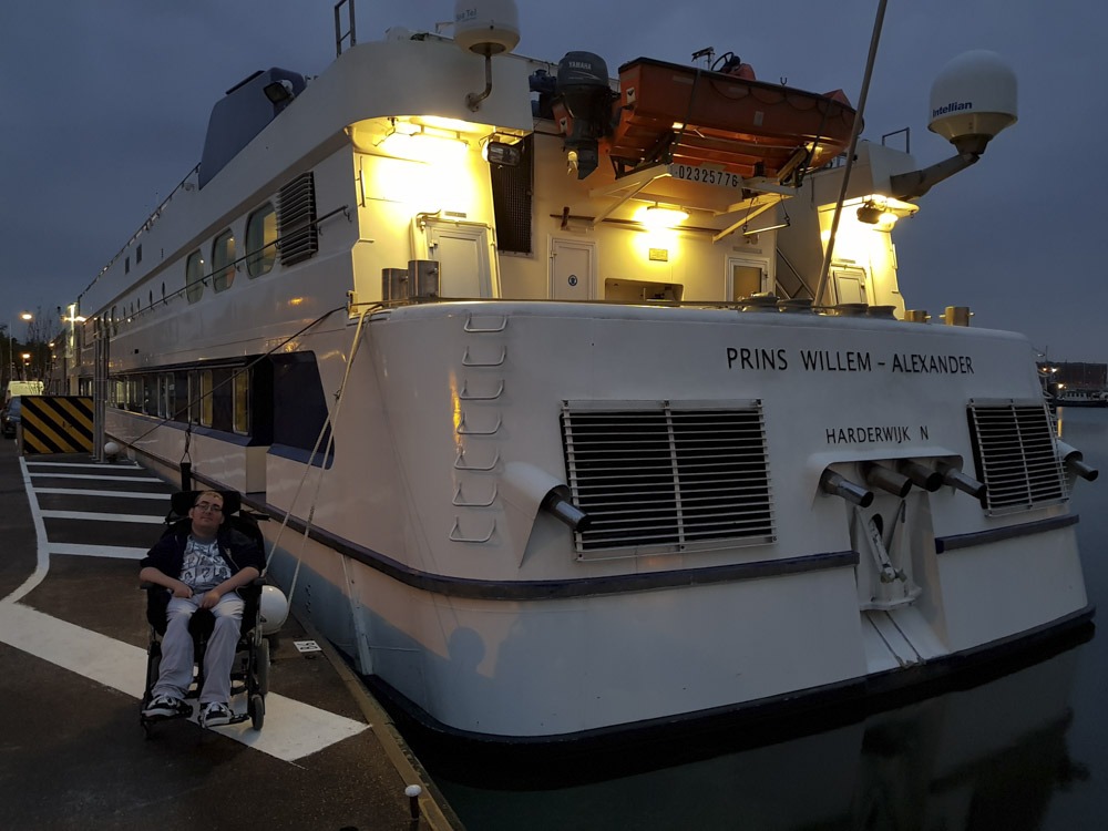 A person in a wheelchair is sitting near the 'Prins Willem Alexander' canal cruise ship, illuminated by lamps, at dusk.