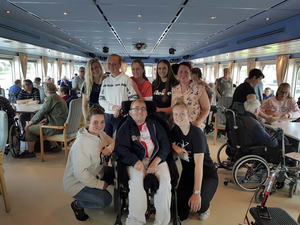 A group of people, including several in wheelchairs, gathered in a well-lit indoor area of a ship, with tables and chairs visible in the background.