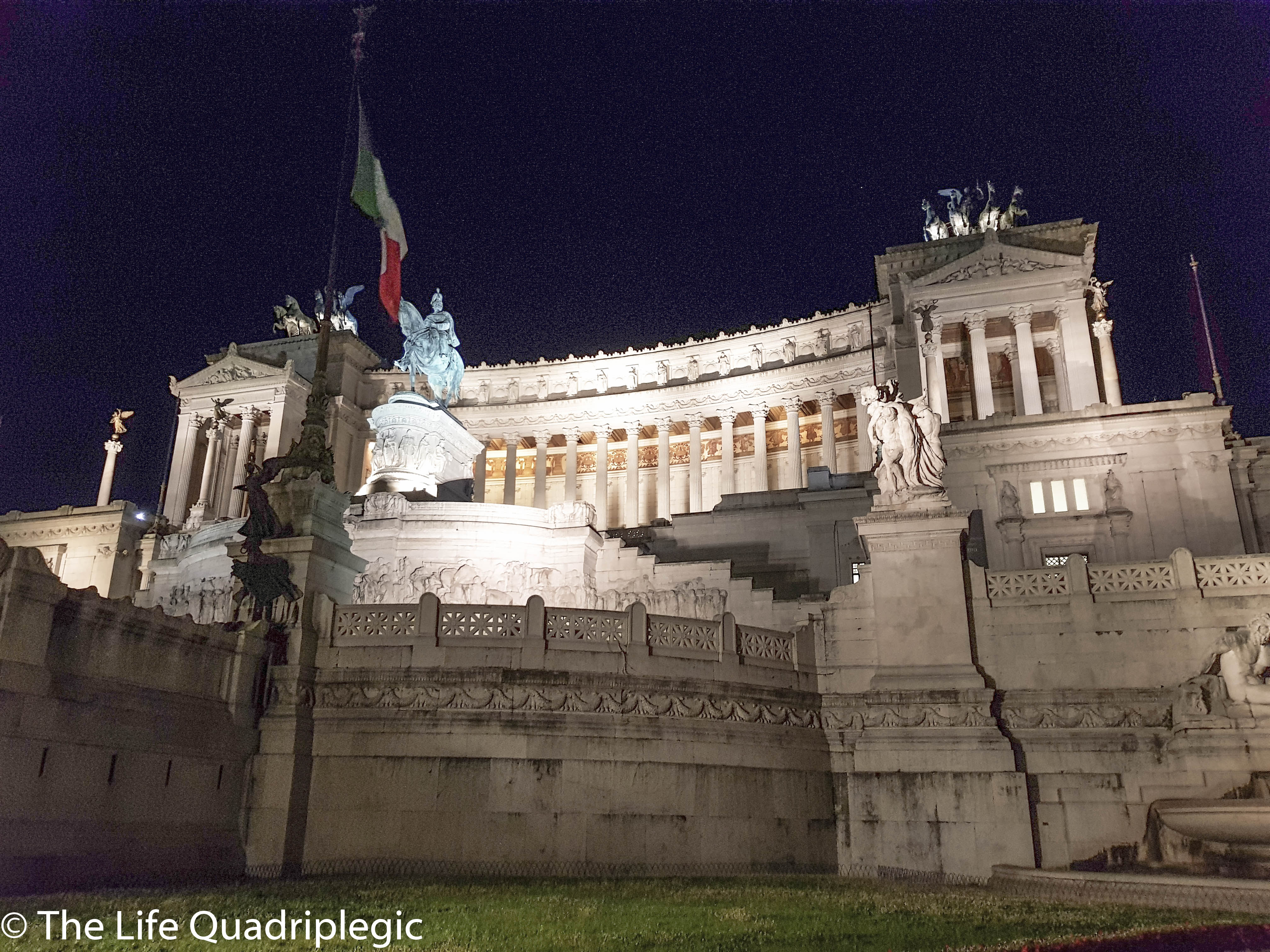 Illuminated view of the Vittoriano Monument in Rome at night, showcasing its grand marble architecture and statues, with the Italian flag flying above.