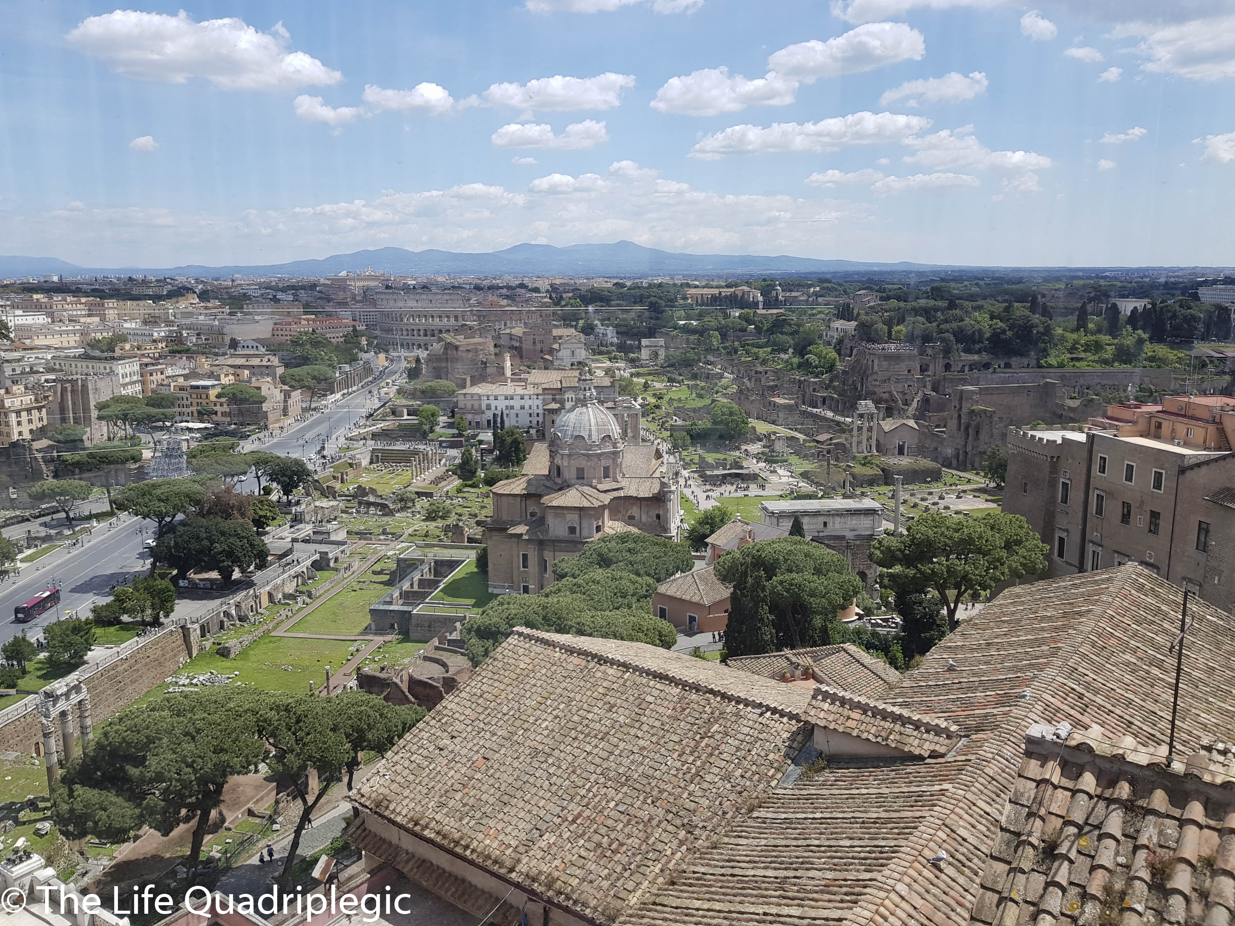 A panoramic view of Rome featuring the Roman Forum and surrounding historic architecture, with a clear blue sky dotted with clouds.