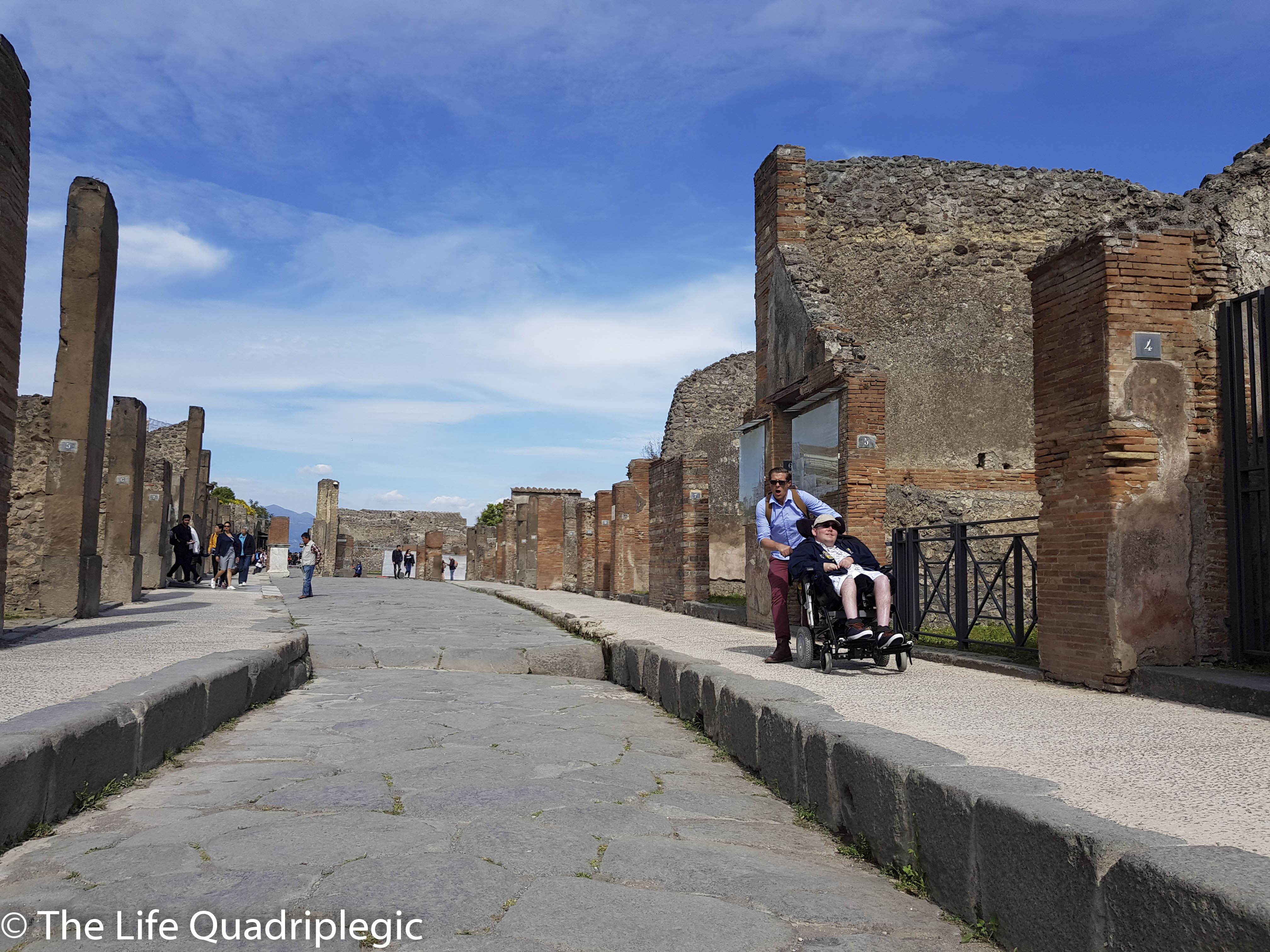 A wheelchair user being pushed by a companion along a smooth pavement at the side of a street in the archaeological site of Pompeii, with ancient ruins and a clear blue sky in the background.