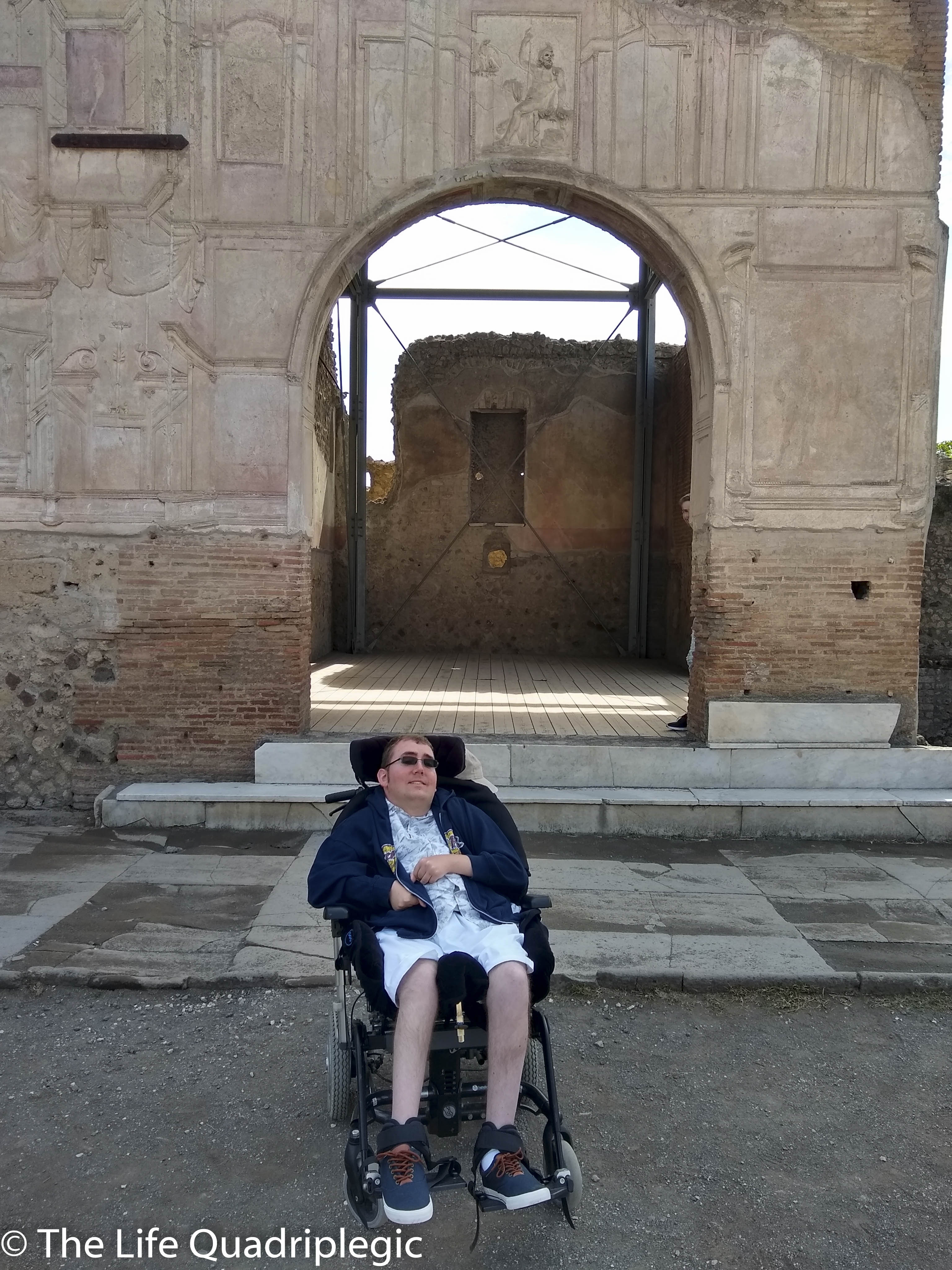 A person in a wheelchair relaxing in front of an ancient archway with faded frescoes and brick walls at the archaeological site of Pompeii.
