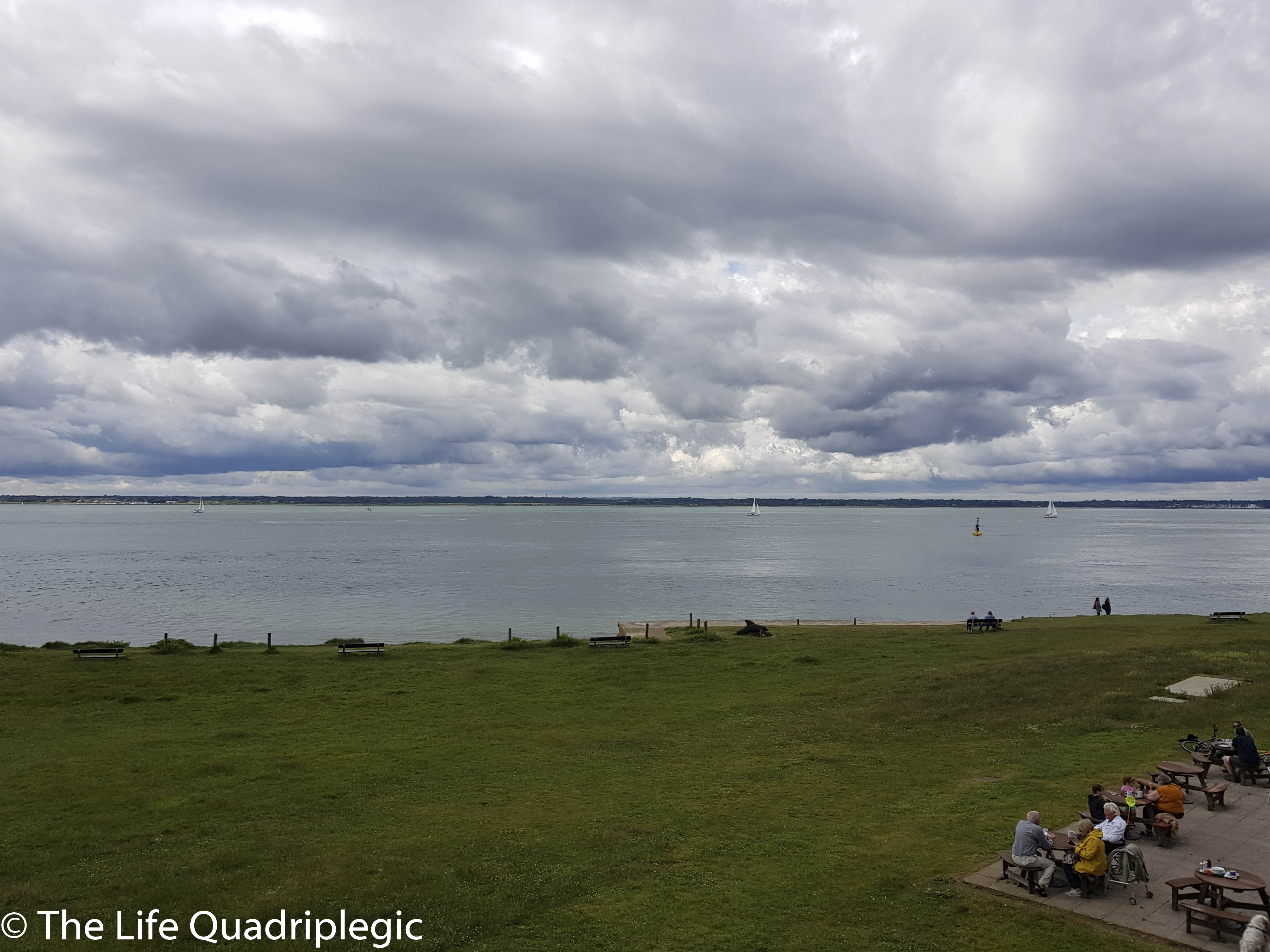 A scenic view of the Solent with sailing boats in the distance and a grassy foreground. The sky is overcast with gray clouds.