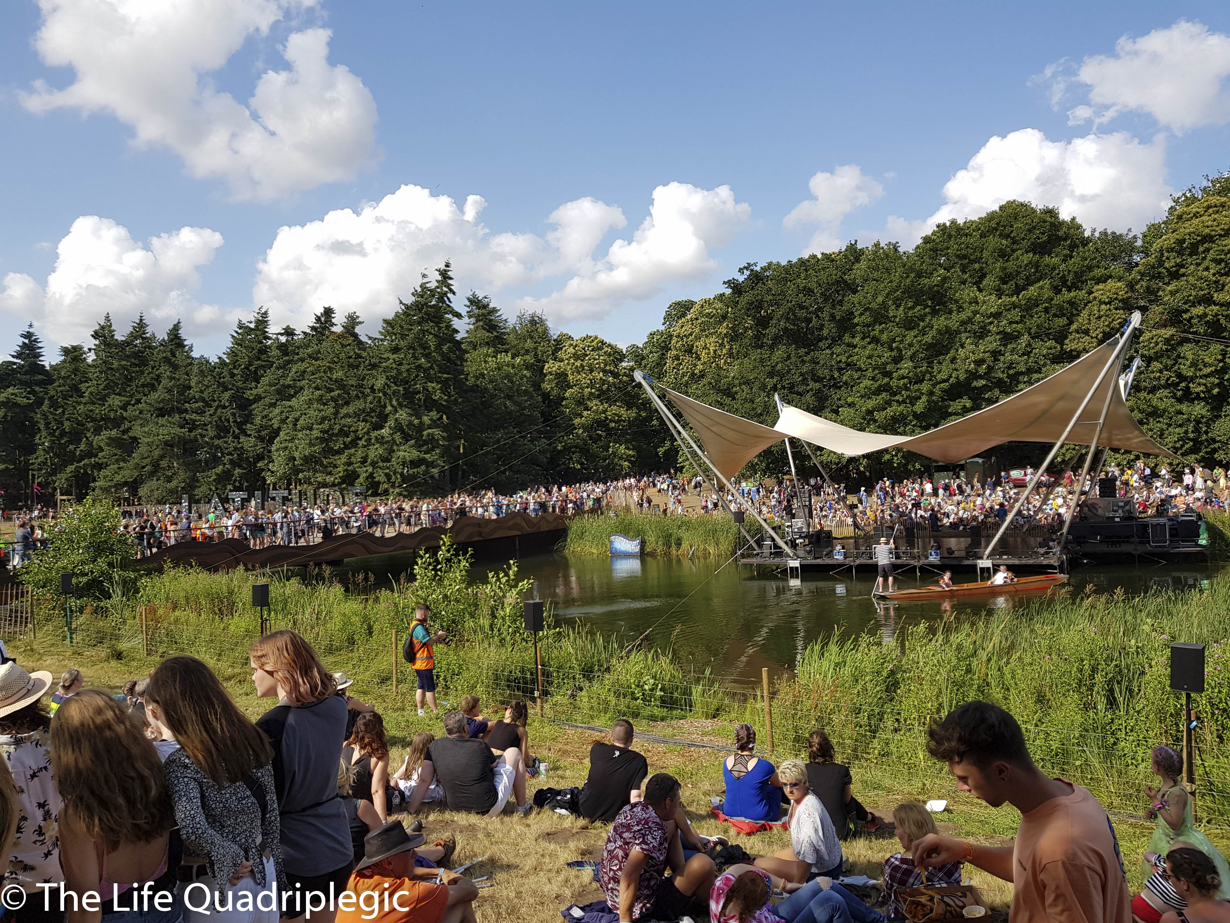 In the foreground people relax on the grass next to a lake. A stage is built over the lake and a crowd is assembled on the far shore