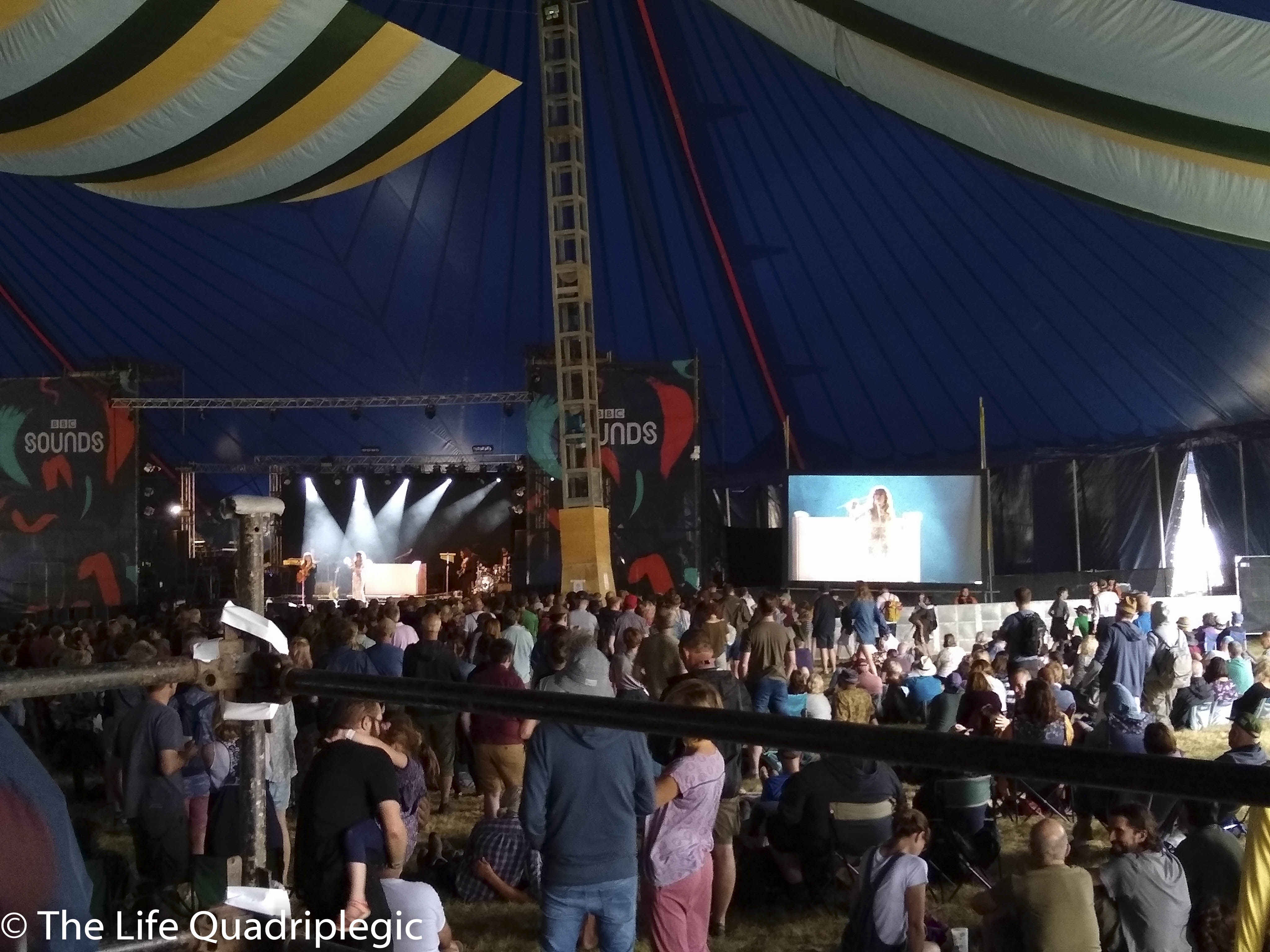 A stage is in the background with a big screen to the right of it. A large crowd is stood in front of the stage watching a band