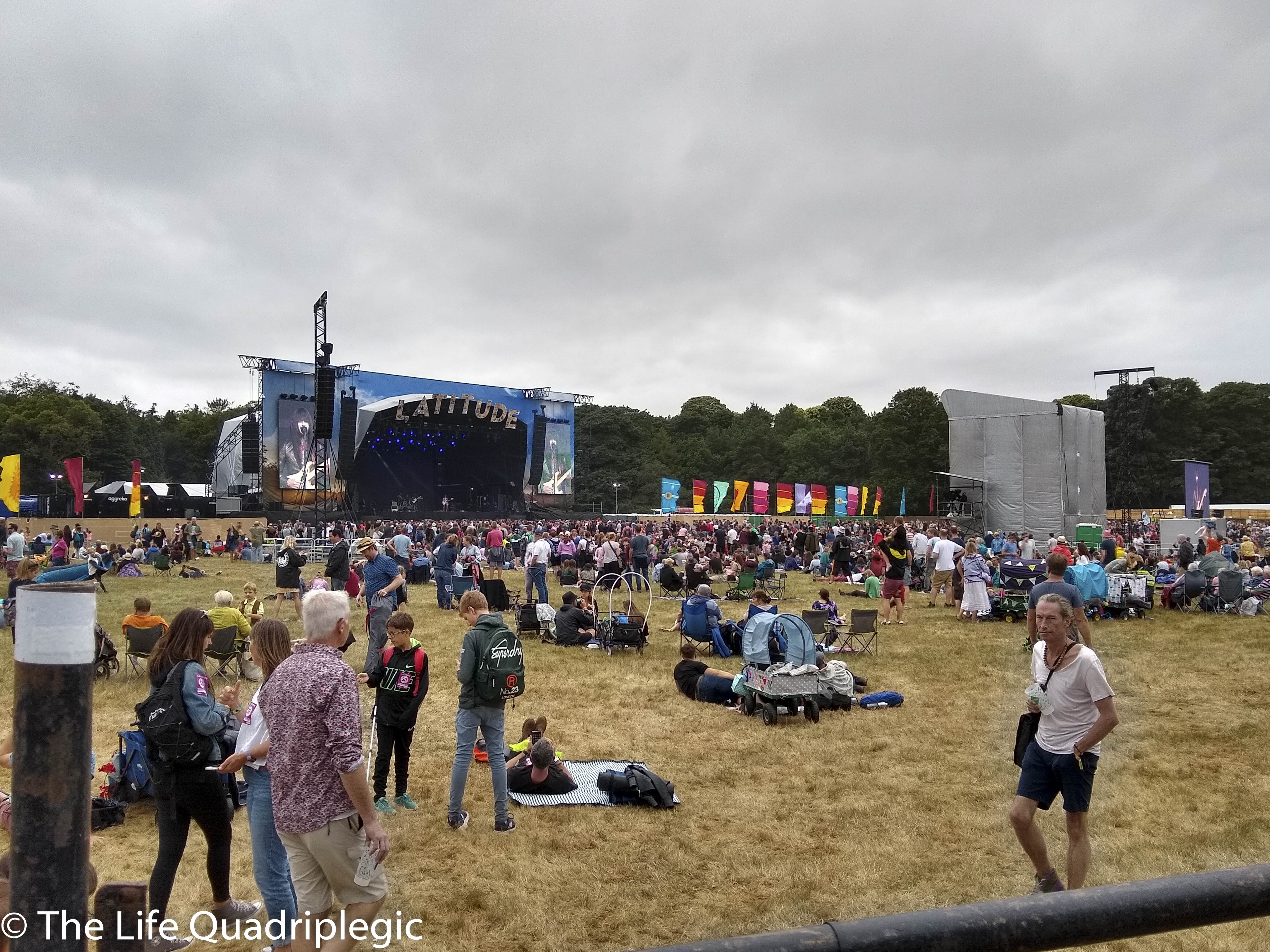 A large stage is in the background with a line of trees behind it. In front of the stage there is a large crowd relaxing in a grassy field