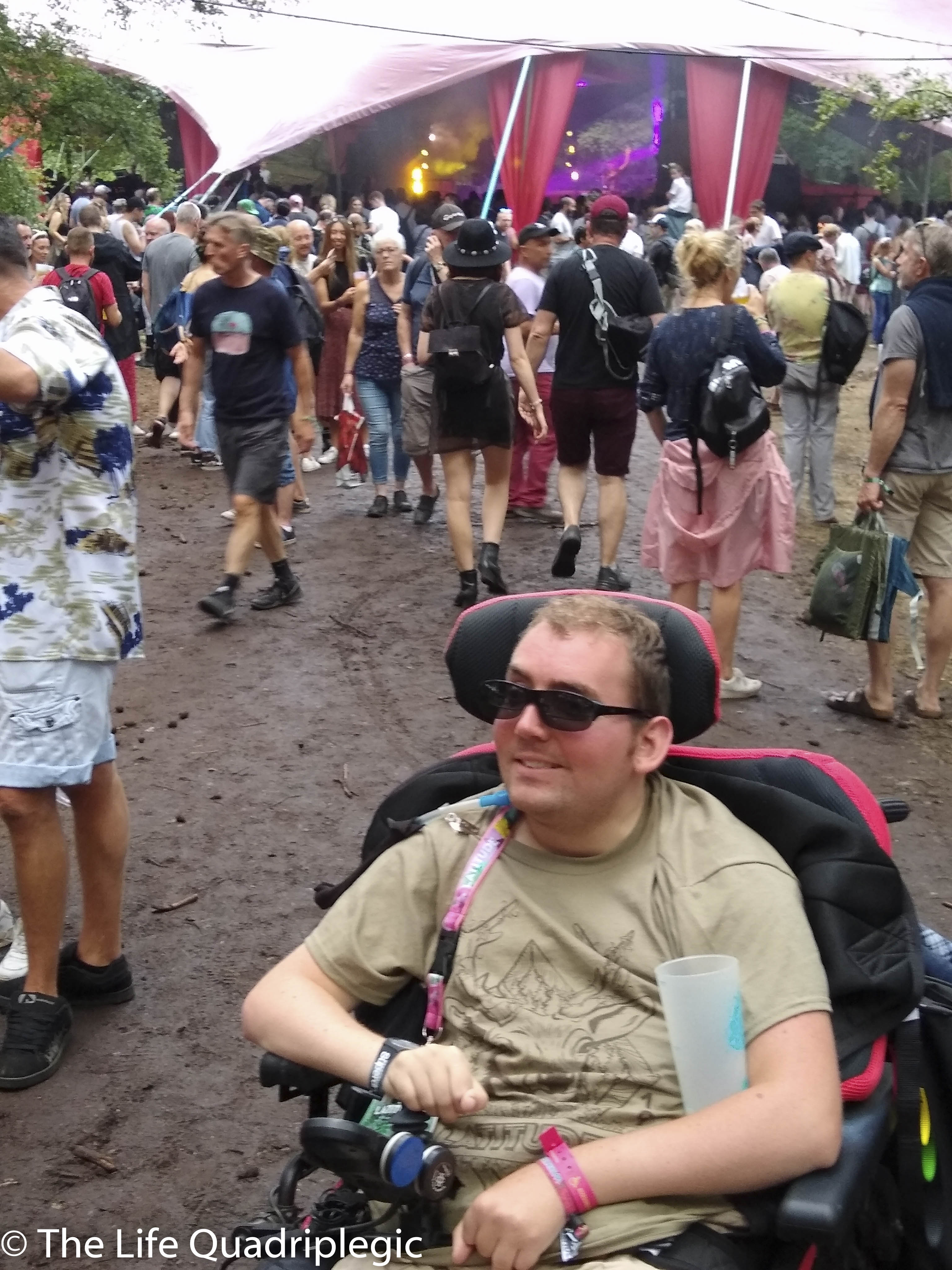 A young man in a powerchair smiles at the camera with a crowd of people walking towards a stage in the background