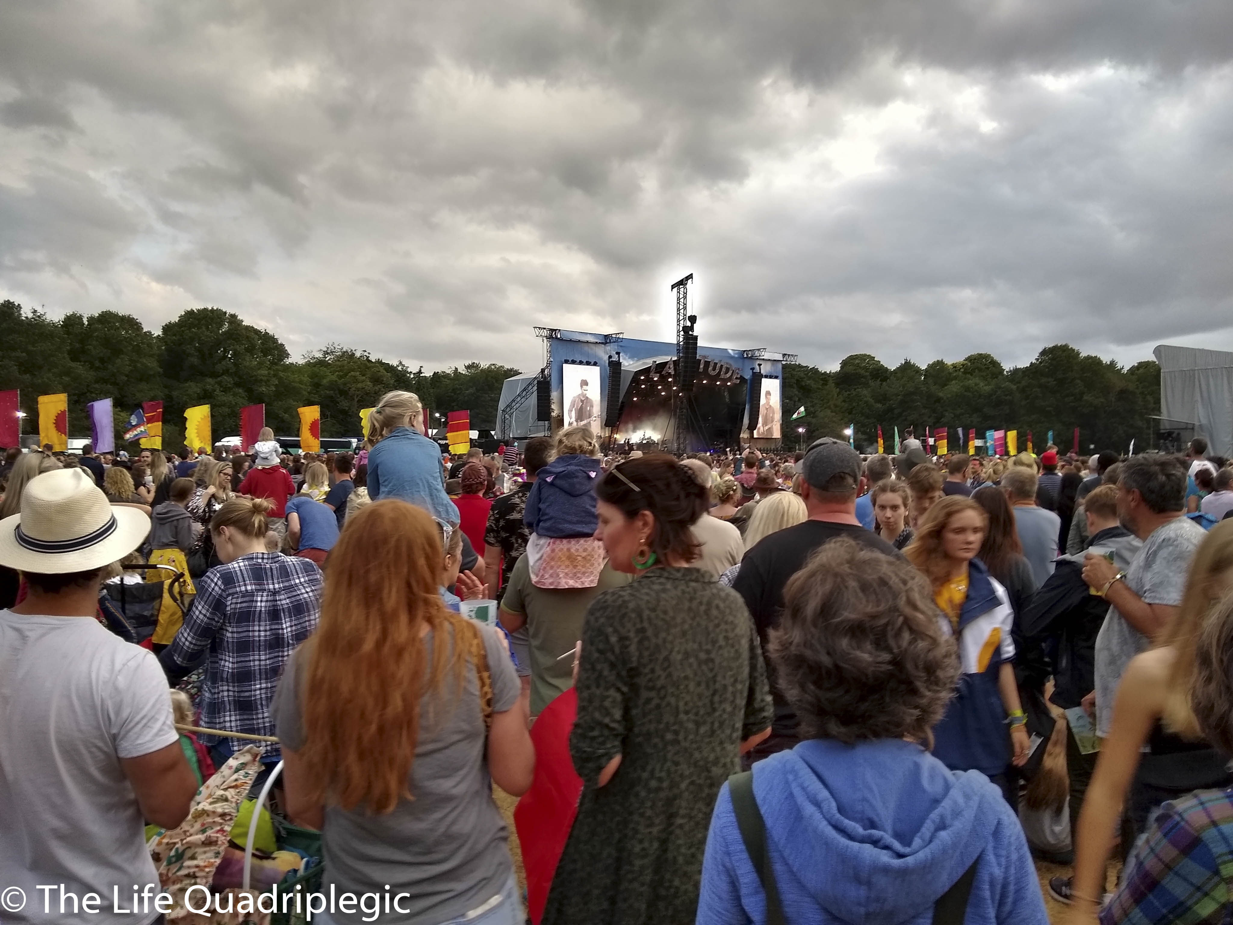 A large crowd is stood in the foreground looking towards a stage in the background under a cloudy sky.
