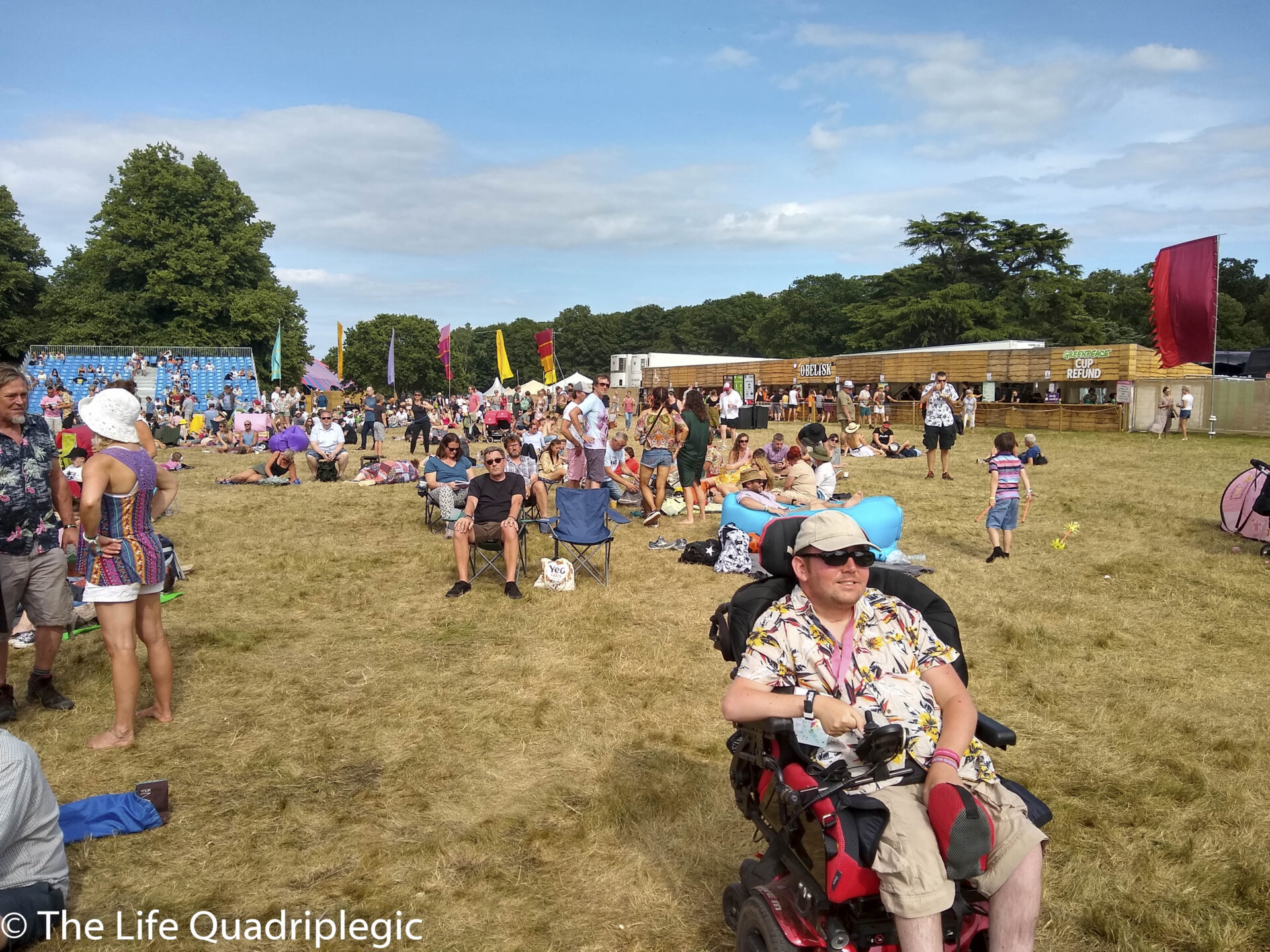 A young man in a powerchair Smiles at the camera while sat in a grassy field. Lots of other people are milling about.