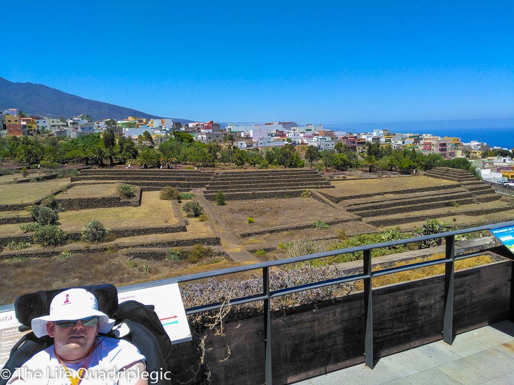 A male wheelchair user sat in front of a railing overlooking stepped pyramids, with some houses beyond.