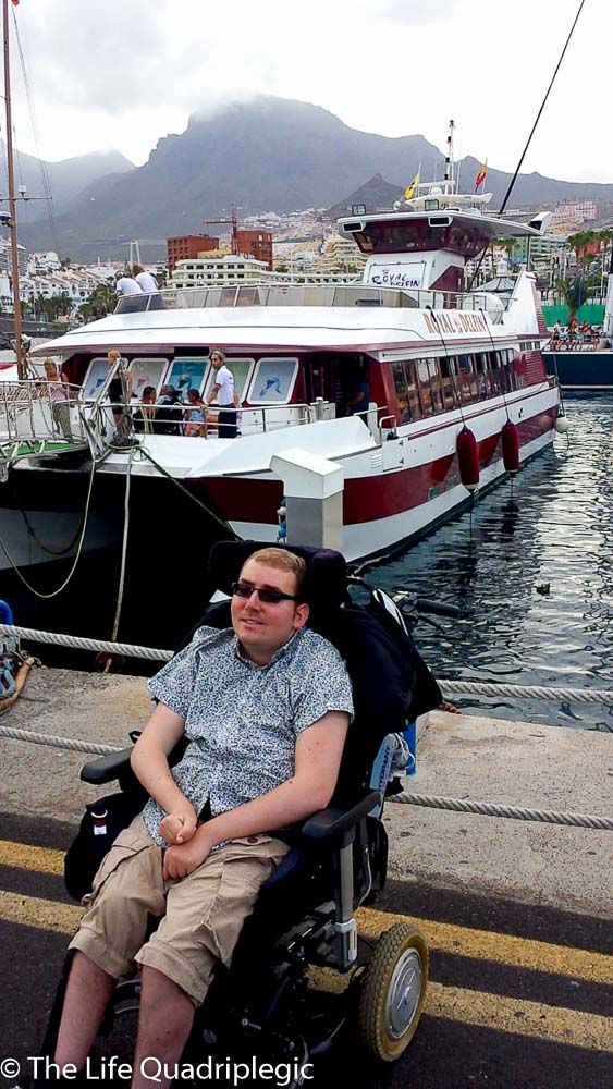 A male wheelchair Use that is that on the edge of the harbour next to the water with a large boat in the background