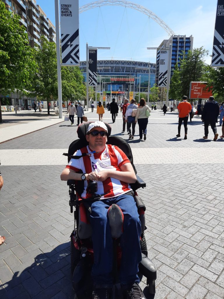 Me sat in my wheelchair on a large flat paved way leading up to Wembley Stadium in the background with several people walking towards it