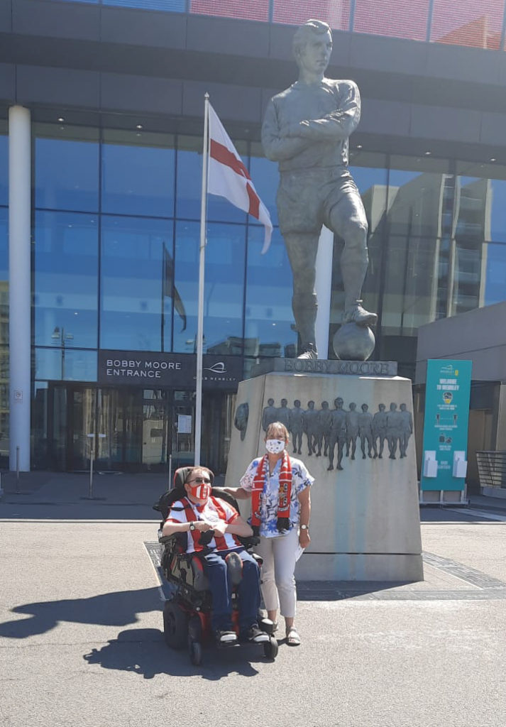 Me and my mum sat in front of a statue of Bobby Moore with his arms folded and 1 foot on a football. The statue stands in front of a big glass windows of the stadium