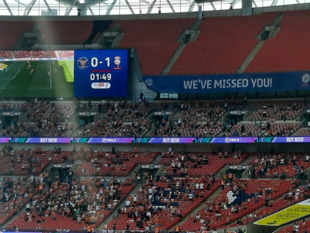 A close-up of a scoreboard which reads Blackpool 0 - 1 Lincoln 1:49. Underneath fans are scattered around the Stands