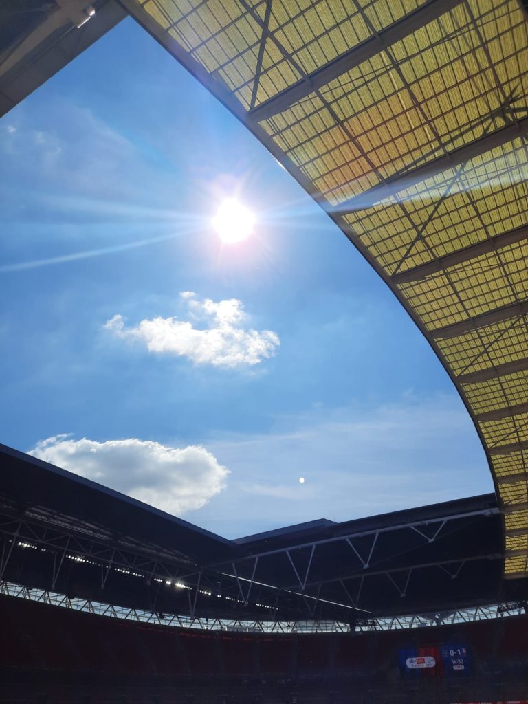 Blue sky with 2 clouds and the sun shining through the open roof of the stadium