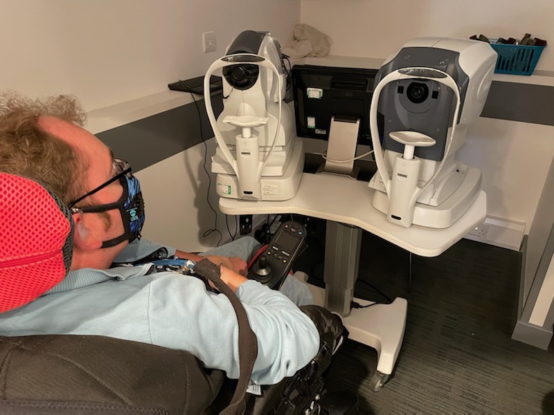 sits in front of a table with 2 machines on it. The machines have a chin rest just below a circular black plans
