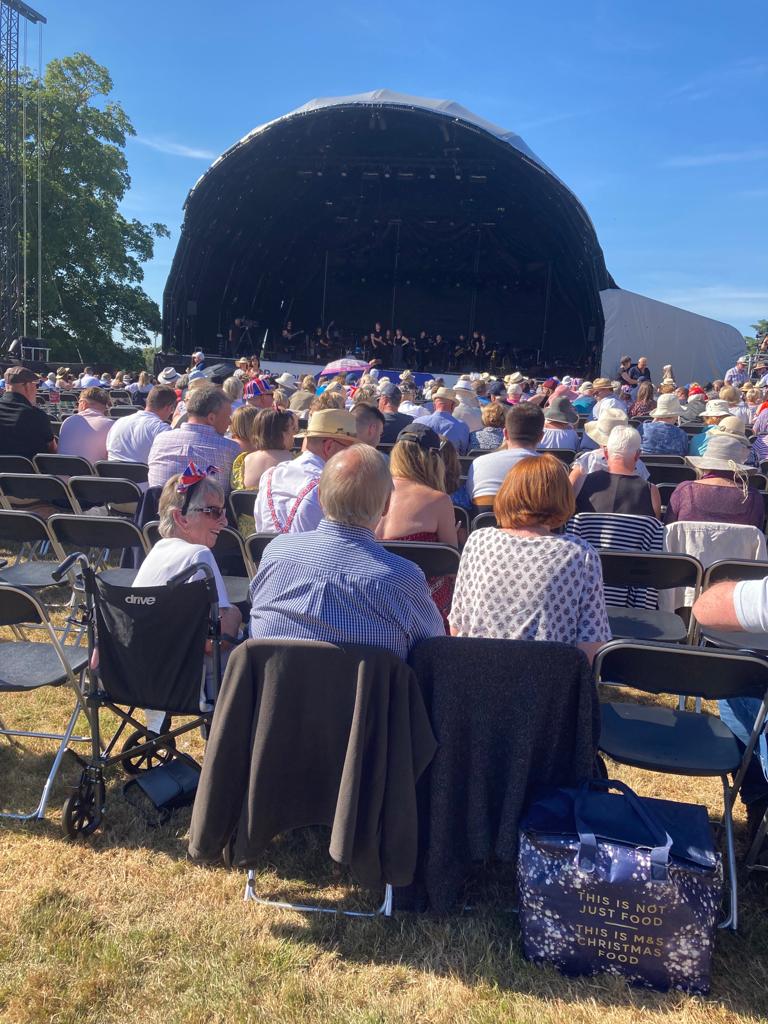 Audience seated in front of a large stage at Leeds Castle concert, with a clear blue sky above and trees in the background.