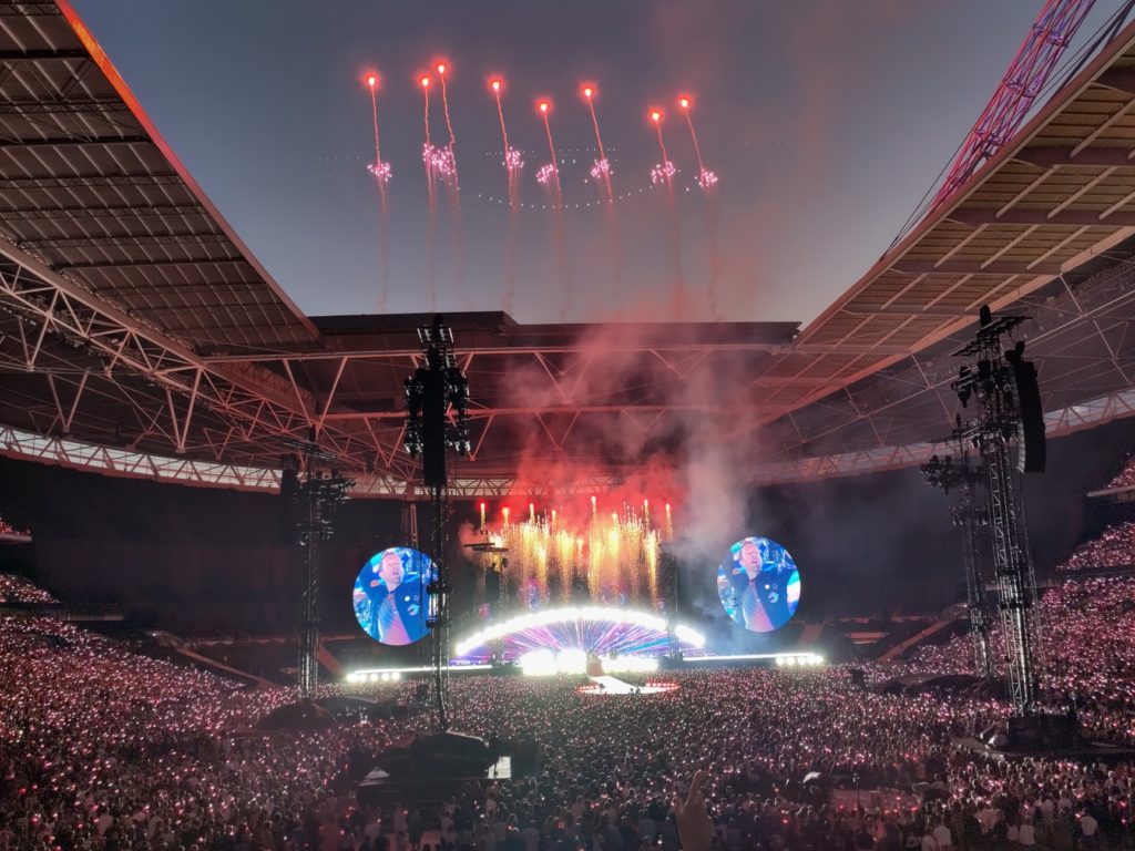 Fireworks shoot up in the sky through the open stadium roof with the stage underneath And large crowds in front of it