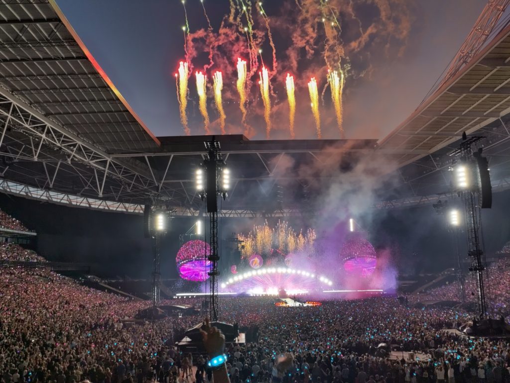 A large crowd feels the area in front of the stage and up the side terraces of the stadium. Fireworks Shoot up in the sky through the open stadium roof