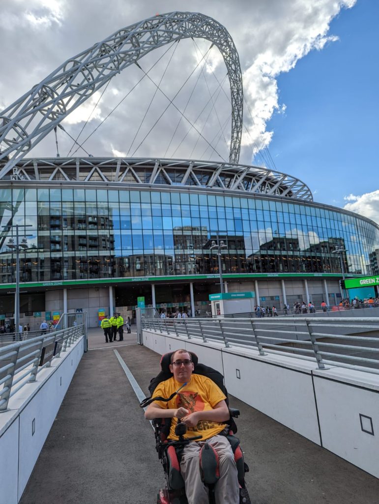 A young man sat in a powerchair facing the camera with Wembley Stadium in the background