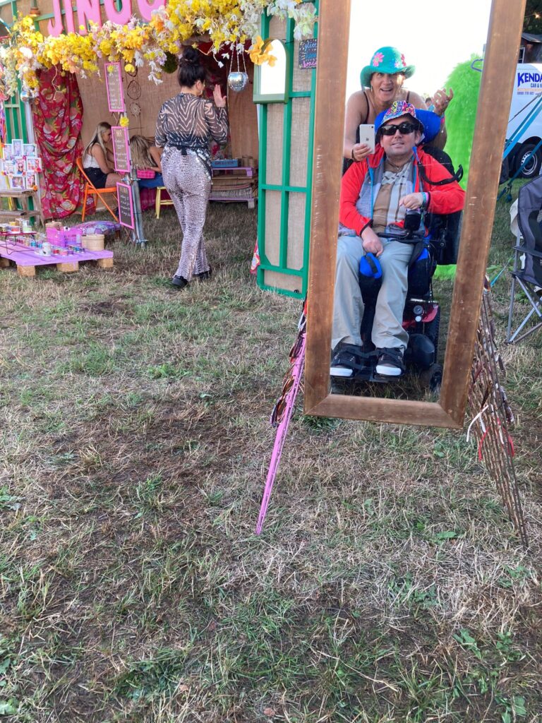 A man in a power chair and a woman wearing colourful hats take a selfie in a mirror with a smart phone