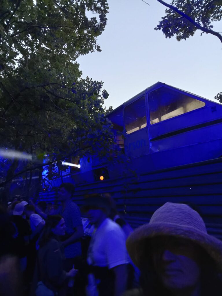 In a forest clearing a disused monorail carriage sits next to people dancing with trees overhead