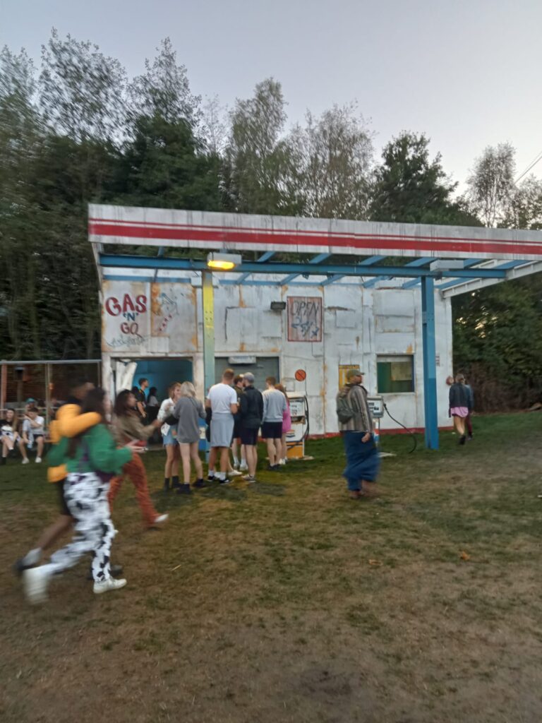 A rundown looking petrol station on open grassy area with trees behind it and people hanging around its pumps