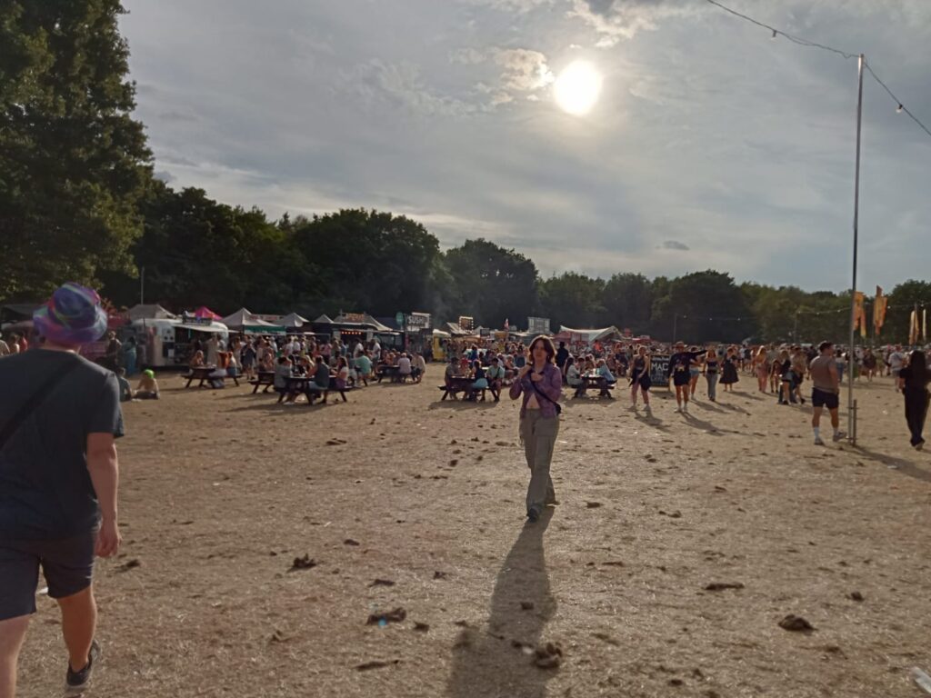 A large number of people congregate around food trucks in a background on a flat grassy area with trees behind the food trucks