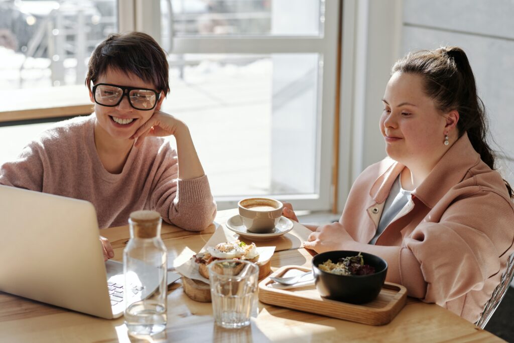 2 young ladies, one with Down syndrome, sit at a table looking at a laptop
