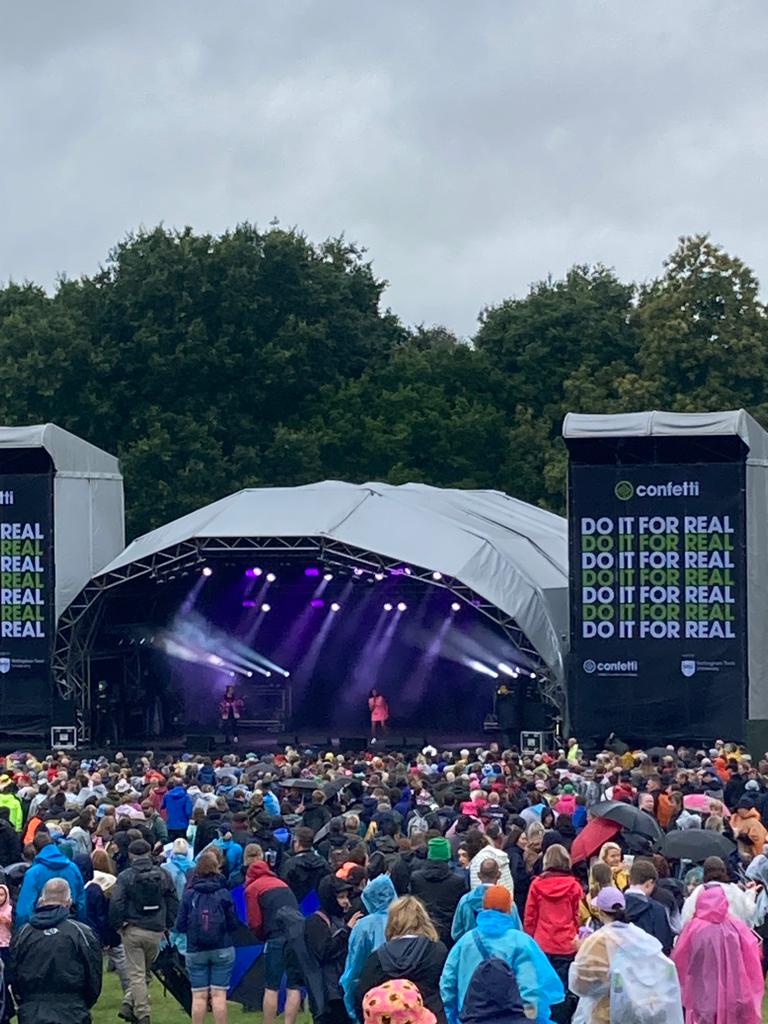 A crowd at Splendour Festival watching a performance on stage, with overcast skies and attendees wearing raincoats and holding umbrellas.