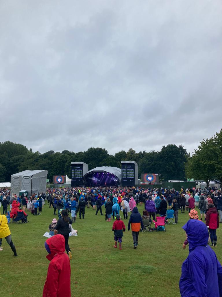 Crowds wearing waterproof clothing gathered under cloudy skies at the Splendour Festival, with a stage and screens visible in the background.