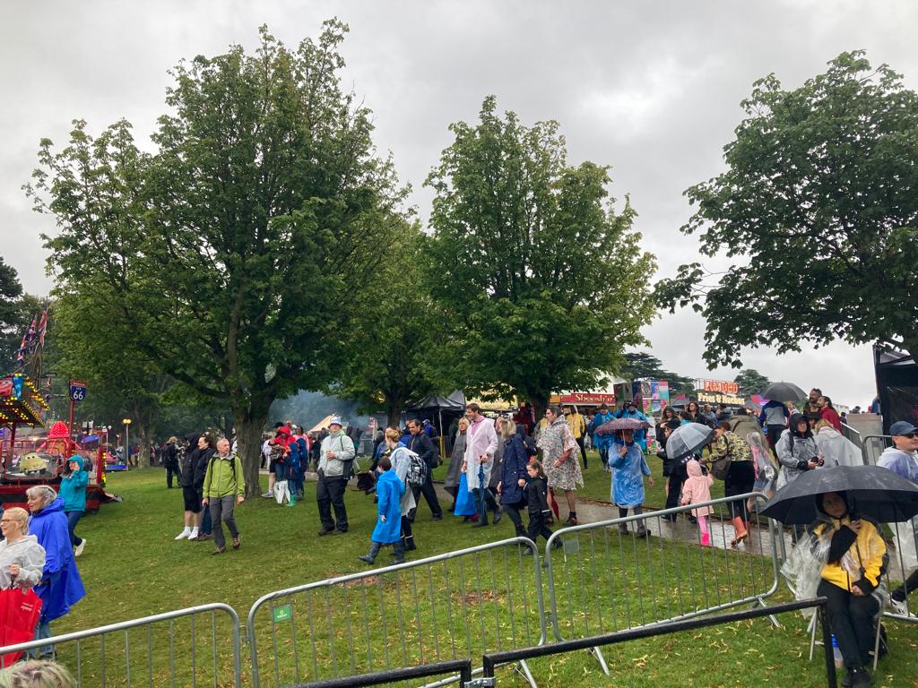 A crowded area at Splendour Festival, with attendees in rain gear and umbrellas on a cloudy day. The scene includes trees and food vendors in the background.