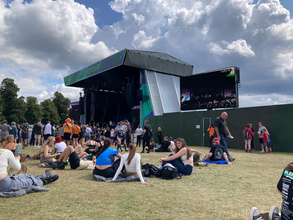 A lively scene at Splendour Festival with a stage in the background, crowds of festival-goers sitting on the grass, and a large screen displaying the performance.