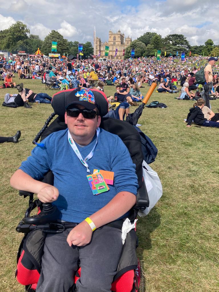 A person in a wheelchair smiles at the camera, surrounded by a large crowd at the Splendour Festival, with Wollaton Hall in the background.