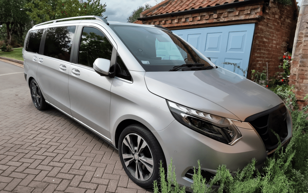 A silver Mercedes V250 wheelchair accessible vehicle parked in front of a house with a blue garage door, surrounded by greenery.