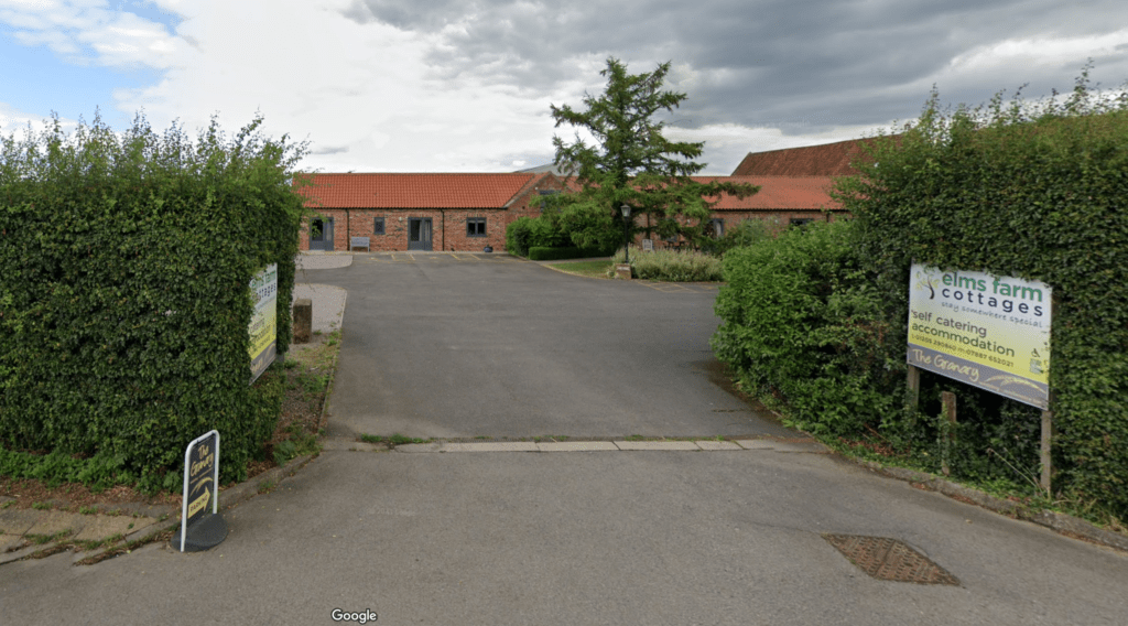 Entrance to Elms Farm Cottages featuring a driveway surrounded by greenery and signage for self-catering accommodation.