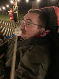 A man in a wheelchair enjoying a giant marshmallow on a stick, sitting in a festive outdoor setting with warm lights in the background.